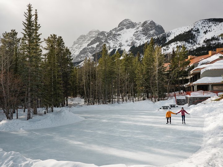 A couple holding hands while ice skating on an outdoor rink , surrounded by trees and mountains.