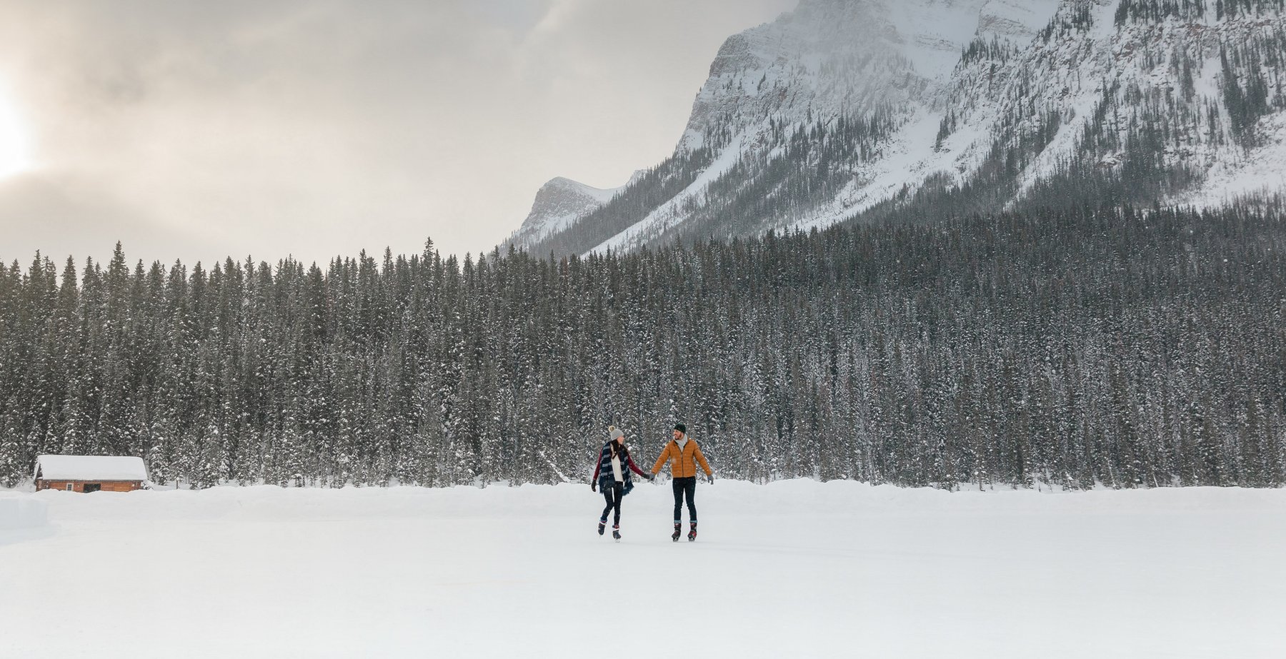 A couple holding hands while ice skating on Lake Louise, mountains and trees in the background.