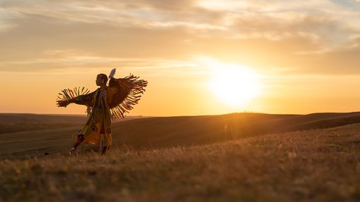 An indigenous dancer performing in front of a sunset among the rolling hills of the prairies.