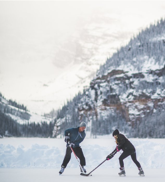 Two people playing ice hockey on a frozen lake with mountains in the background.