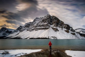 A winter hiker in a red jacket pauses to view an imposing mountain and Bow Lake in Banff National Park under a dramatic blue-grey sky.