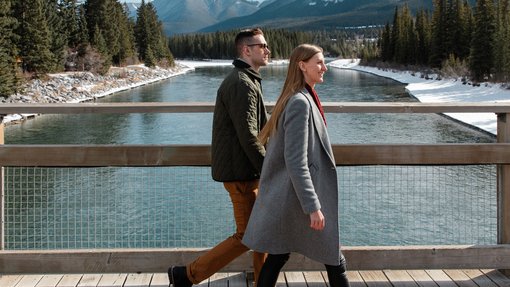 Couple walking along a riverside bridge with mountains in the background