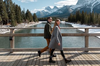 Couple walking along a riverside bridge with mountains in the background