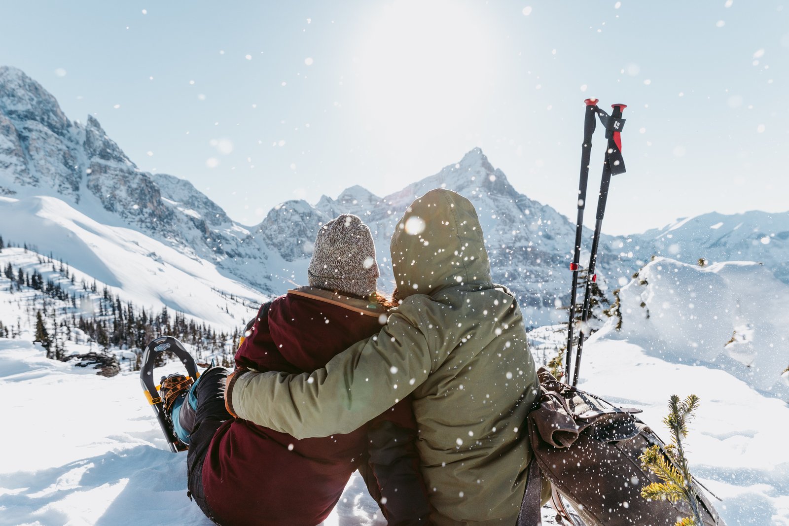 Couple in a sitting embrace wearing snowshoes and looking out at the mountains covered with snow.