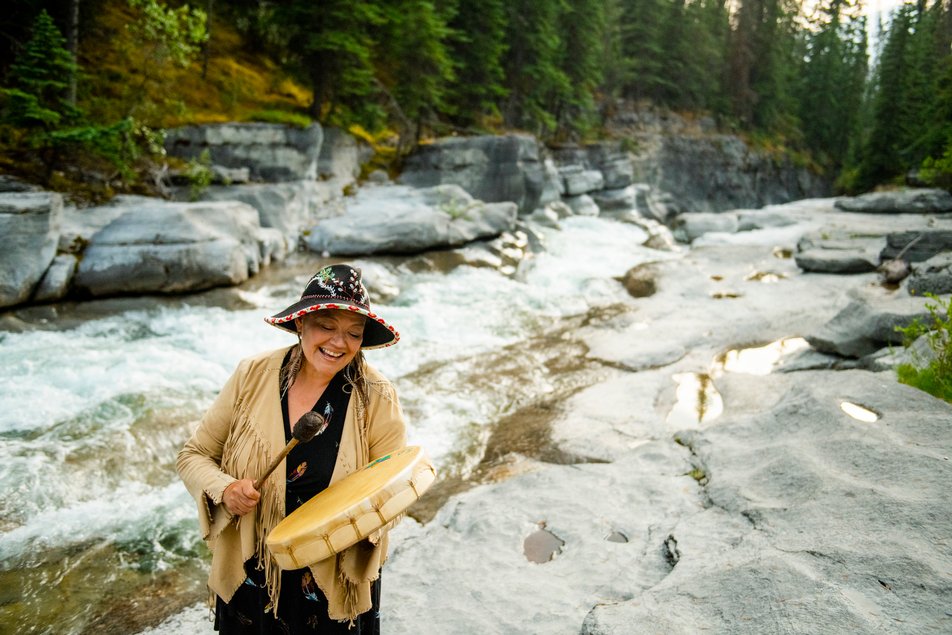 Female Indigenous cree elder from Warrior Women plays a hand drum in front of a river in Jasper National Park.