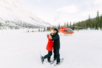 A snowshoe-clad couple hugs in front of a helicopter near Marvel Pass in Banff National Park.