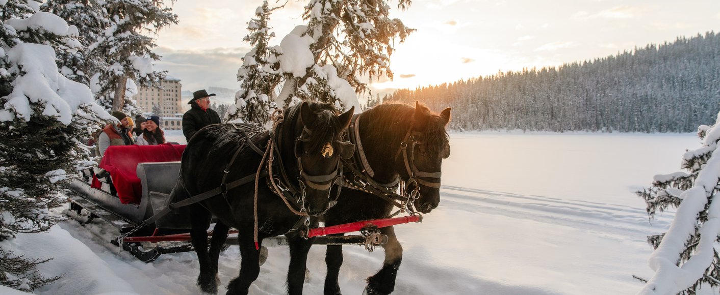 The sun rises behind a sleigh ride along the snowy path beside frozen Lake Louise on an idyllic winter day in Banff National Park.