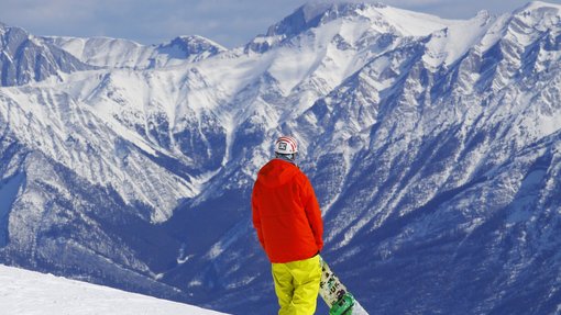 A snowboarder holding their board while looking at the mountain view from the top of a ski resort run.