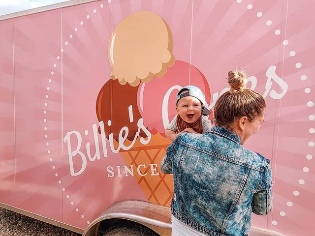 A woman holding a baby standing in front of the pink Billie's Cones ice cream truck.