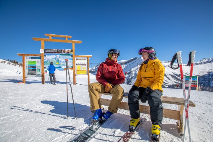 Two skiers sit on a bench at the top of a run.