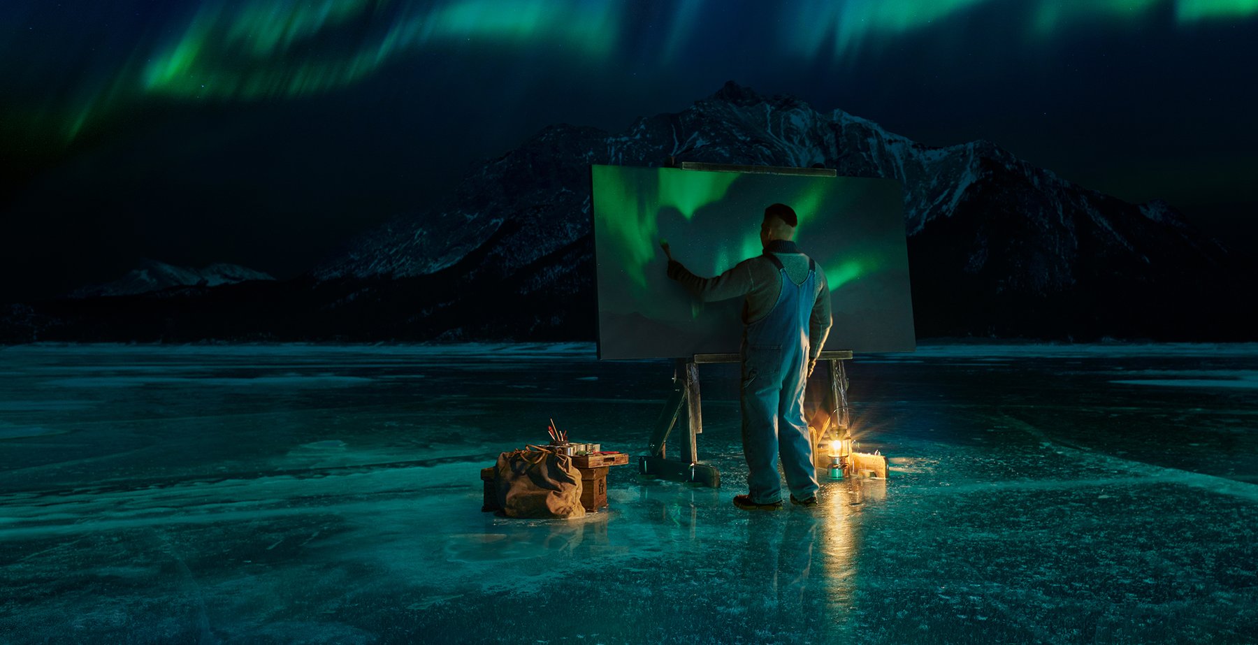 Man standing on frozen lake at night painting the Northern Lights on a large canvas with the Northern Lights overhead in the sky.
