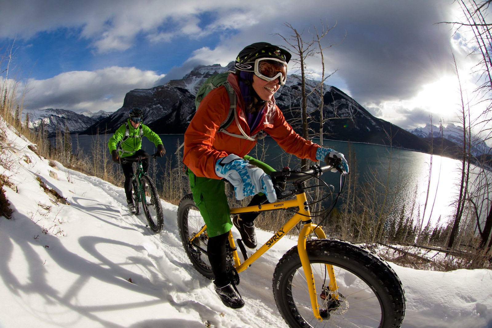 A woman and a man smiling as they ride fat bikes along a snowy trail beside a mountain lake.