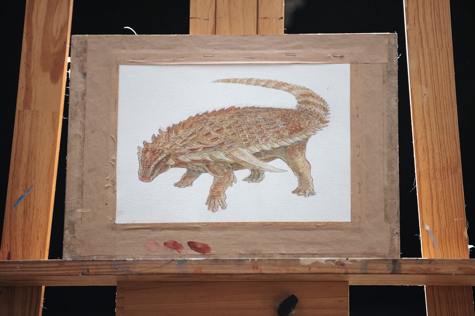 A painting of a Borealopelta markmitchelli sits on a wooden easel.