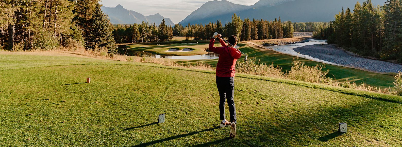 A golfer tees off at the Kananaskis Country Golf Course with mountains in the background.