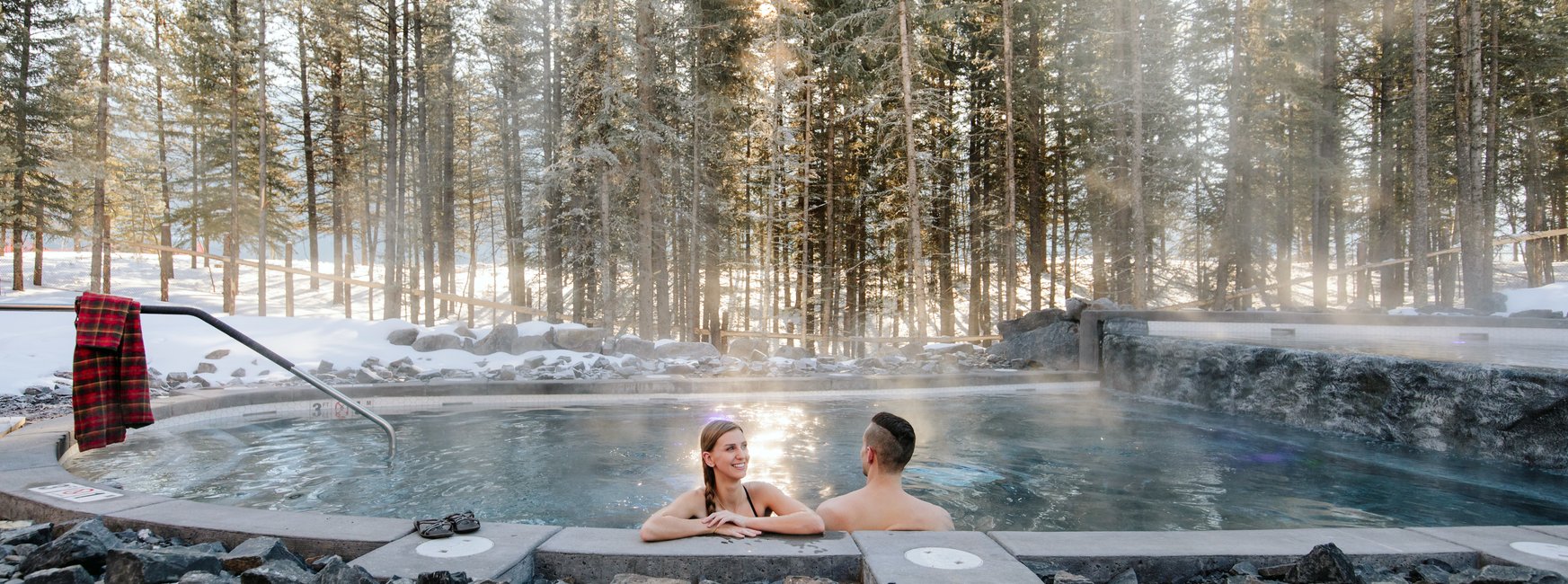 Snow scene with a couple soaking in an outdoor hot pool in the woods. Kananaskis Nordic Spa at the Pomeroy Kananaskis Mountain Lodge. Canadian Rockies.