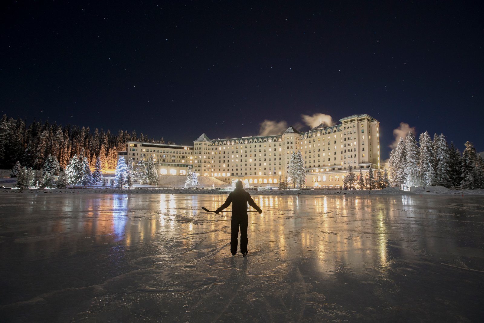 A lone ice skater skating and holding a hockey stick on a frozen lake at night with a lit hotel in the distance.