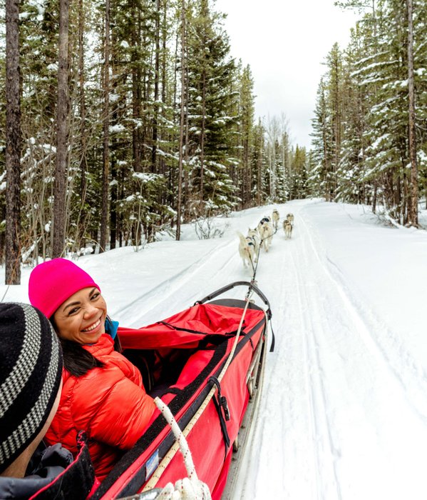 A women participant turns and smiles at the camera while on a dog sledding tour through a snow covered forest.