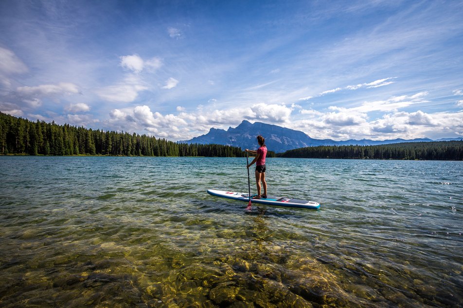 Stand-up paddleboarder paddles on Two Jack Lake with mountain views and trees around the shore.