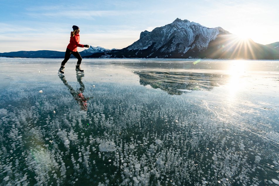 A woman skates across a frozen lake with mountains and sun in the background.