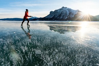 A woman skates across a frozen lake with mountains and sun in the background.
