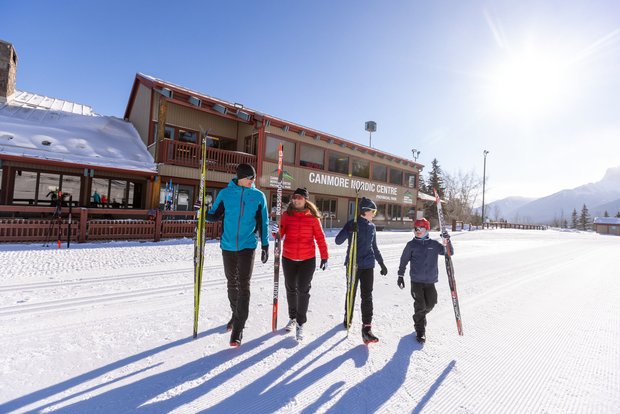 Family of four walking out of the ski lodge with their skis in hand at the Canmore Nordic Centre.