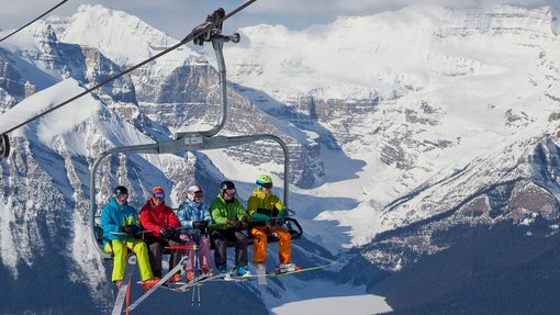 A mix of five skiers and snowboarders riding up a chairlift with mountains in the background.