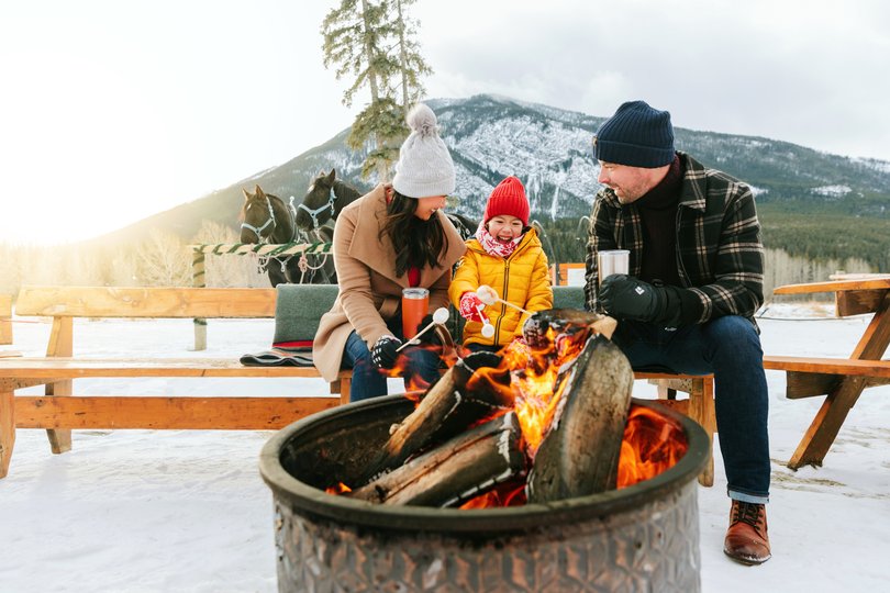 A family roasting marshmallows over a fire after a winter horse-drawn sleigh ride in Banff National Park.