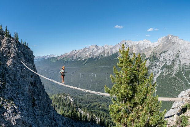 A woman walks along a via ferrata walkway between two mountains above a forested valley below.
